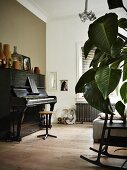 Mit Vasen dekoriertes, altes Klavier gegenüber Sofa; Blätter einer Zimmerpflanze und Schaukelstuhl-Ausschnitt im Vordergrund