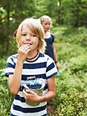 A little boy in a forest eating freshly picked blueberries with a little girl in the background