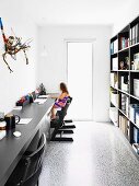 Narrow home office with black furniture and flotsam artwork above long desktop opposite bookcase