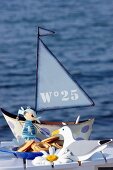 Kindliche Dekofiguren und Gebäckschale vor Miniatur Segelboot im Hintergrund Wasser