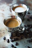 An espresso and a cappucino on a metal surface with coffee beans