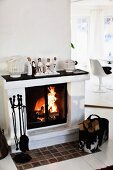 Fireplace with festively decorated, traditional mantelpiece, classic fire irons and bag of firewood; modern dining area in background