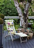 Rustic wooden lounger with cushions next to urns and tree on dark grey wooden deck