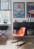 Charles Eames rocking chair with orange shell seat in front of floating sideboard below framed posters