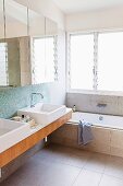 Modern bathroom with mirrored cabinet above twin sinks next to bathtub below window