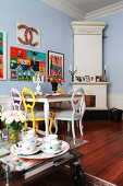 Dining area with antique chairs painted different colours and bright artworks on wall in period apartment; glass table on castors in foreground
