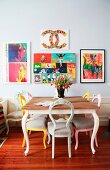Dining area with antique chairs painted in various colours and colourful pictures on light grey wall in period apartment