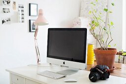 Computer und Klemmlampe auf weißem Schreibtisch, dahinter Zimmerpflanze im Tontopf