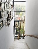 Narrow stairwell with framed pictures on wall, floor-to-ceiling glass facade at foot of staircase with view of terrace