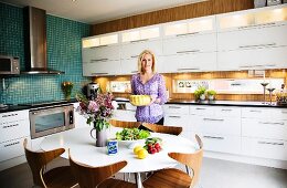 Fitted kitchen with white cupboard doors, sections of wood panelling on wall, white dining table and shell chairs