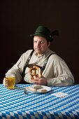 A stereotypical German man wearing lederhosen and eating a pretzel and white sausage with beer