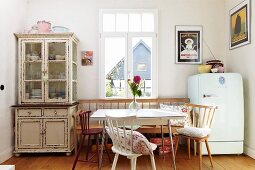 Dining area in rustic ambiance with chairs and bench below window between dresser with glass-fronted top cabinet and fridge