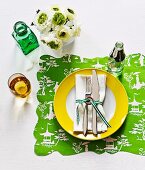 Cutlery and napkin tied with ribbon on plate with yellow edge and place mat made from printed wrapping paper with wavy edge