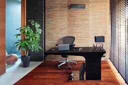 Modern, black desk with projecting top, louvre blinds on window and house plant next to glass partition in study