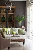 Toile de jouy ottoman in seating area with French style emphasised by lantern pendant lamp and demijohn bottles in wooden, glass-fronted cabinet