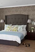 Box spring bed with tall headboard and elegant table lamps on bedside cabinets against brocade-patterned wallpaper