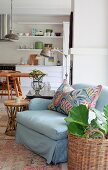 Pale blue armchair with colourful scatter cushion and house plant in wicker basket on floor; dining area in open-plan kitchen in background