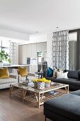 Grey sofa and grey and white striped armchairs around coffee table made from wooden slats with glass top; open-plan kitchen with fitted cupboards in background