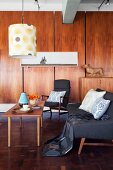 Sofa and armchair with grey upholstery around simple wooden coffee table below pendant lamp with retro-patterned lampshade; fitted wooden cupboards in background