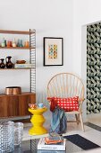 Yellow, zigzag stool and chair with high, curved backrest and radiating struts next to shelving unit with fifties-style base cabinet