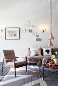 Fifties Sessel und graues Sofa um Couchtisch unter Pendelleuchten mit kupferfarbenem Schirm in loftartigem Ambiente mit Wandtellern auf geweisselter Ziegelwand