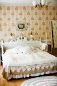 Double bed with white, carved headboard against wallpapered wall in traditional bedroom in shades of white and pale brown