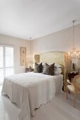 Elegant bedroom; white bedspread on double bed with tall headboard upholstered in glossy fabric next to chandelier