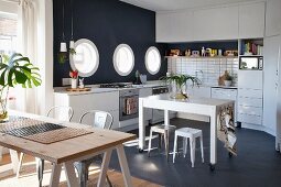 Table on castors in white fitted kitchen with black mosaic floor, black accent wall, porthole windows and wooden table on trestles in foreground