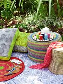 Pouffe, raffia stool and floral floor cushions on picnic blanket in garden