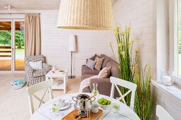 White dining table and chairs below rattan lampshade in front of pale grey sofa set in corner of wood-panelled interior