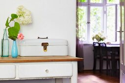 Vintage bread box and vases of flowers on console table with white drawers; view through open door into dining room with table below window