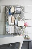 Fabric-covered pin board with grid of diagonal ribbons above grey shabby chic desk and white chair