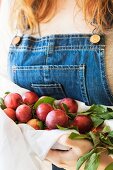 A woman wearing a pair of denim dungarees holding freshly picked red plums in a white cloth