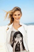 Junge, blonde Frau am Strand mit offener weisser Lederjacke über Motivshirt