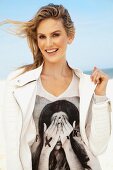 A young blonde woman on a beach wearing an open white leather jacket over a printed T-shirt