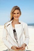 A young blonde woman on a beach wearing an open white leather jacket over a printed T-shirt