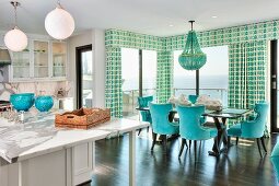 Offene weiße Küche mit türkisfarbener Essecke vor Fensterfront mit Gardinen & Meerblick