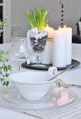 Place setting on dining table; bowl with handle decorated with cord, glass plate, wine glass and arrangement of candles and plants on tray