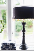 Table lamp with mother-of-pearl sequins decorating black lampshade and turned base in front of window