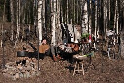 Campfire, picnic, table, tent and bicycle in autumn woods; man relaxing with back against tree trunk