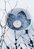 Porzellanschale im Schnee mit Teelicht und ausgeschnittenem blauen Stoffstern mit Silberglitter; unscharfe Zweige im Vordergrund