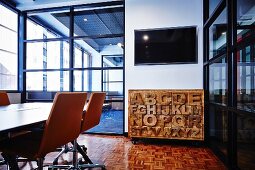 Drehstühle mit hellbraunem Lederbezug an Tisch, im Hintergrund Kommode aus Holz mit geschnitzten Buchstaben neben raumhohem Fenstersprossenelement und Blick ins Nebenzimmer