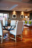 Various vases on round glass table and white upholstered chairs; easy chairs under ceiling spotlights in corner of grey-painted room