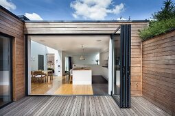 Wooden terrace of contemporary house with wood-clad facade; view through open folding door into kitchen