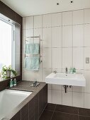 Wall-mounted washbasin on white, vertical tiles and bathtub integrated into black element below window in designer bathroom