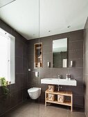 Grey-tiled bathroom with shelves in niche above toilet next to sink and wooden shelves; glass partition in foreground