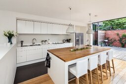 Bar stool with white loose covers at island counter in open-plan designer kitchen with open terrace doors to one side