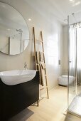 Trough-style sink with dark wood base cabinet, oval mirror on tiled wall, ladder used as towel rack and partially visible glass shower cabinet
