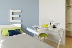 Single bed with headboard and large ornamental letter on pastel blue wall; white metal table and chair against partition wall