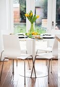 White wooden chairs at a round table laid with a bunch of yellow flowers with garden doors in the background